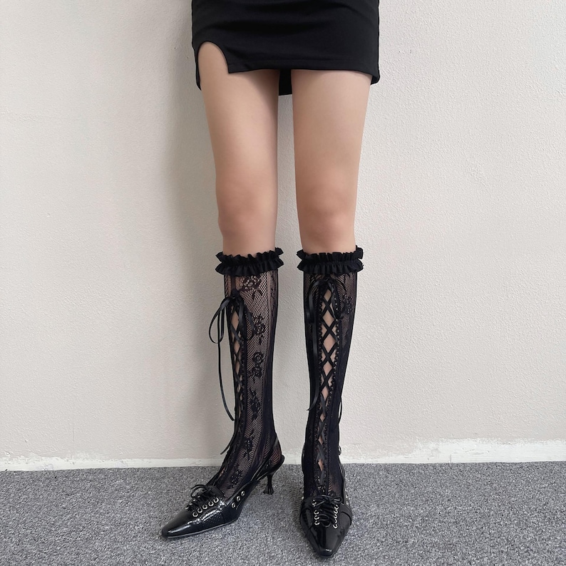 May include: Black lace knee-high socks with a floral design and ribbon ties, worn with black patent leather heeled shoes. The socks feature a ruffled top edge.