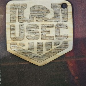 Op de afbeelding: Een lichtgekleurd houten schildvormig label met een lasergegraveerd ontwerp. Het bovenste gedeelte toont een gestileerd berensilhouet boven de tekst USEC, met een geometrisch chevronpatroon aan de onderkant. Rechtsboven bevindt zich een klein gaatje.