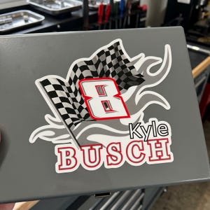 May include: A gray metal box with a sticker featuring a checkered flag and the number 8 in red and white. The name "Kyle Busch" is printed below the number in red letters.