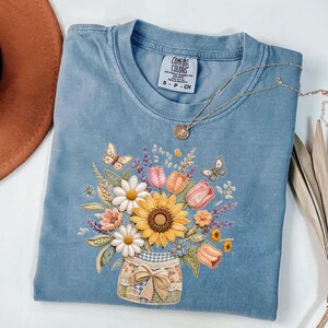 Puede incluir: Una camiseta azul Comfort Colors con un diseño floral bordado. El diseño incluye un girasol, margaritas, tulipanes y mariposas dispuestas en un tarro. También se ve un collar plateado con un colgante.