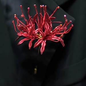 May include: A red spider lily lapel pin featuring long, slender, curved petals and thin stamens tipped with small beads. The flower is attached to a metal pin stem and is positioned against a solid black fabric background.