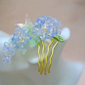 May include: A hair comb featuring a cluster of translucent light blue hydrangea flowers with small white centers and green leaves. The decorative floral arrangement is attached to a gold-toned metal comb with four wavy prongs for securing hair.