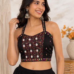 May include: A black halter-neck crop top featuring a central panel with a colorful geometric diamond pattern, white cowrie shell embellishments, and thin straps. The garment includes intricate multi-colored embroidery along the edges and neckline.