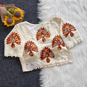May include: An off-white blouse featuring short sleeves and a repeating pattern of orange and brown floral-like motifs. The garment is finished with a white pom-pom trim along the hem and sleeve edges, and includes a small white tag labeled VVI.