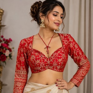 May include: A red long-sleeved blouse featuring a square neckline and an intricate gold-toned geometric pattern throughout. The garment is paired with a cream-coloured skirt and is accessorised with gold-toned dangling earrings and a red cord necklace.