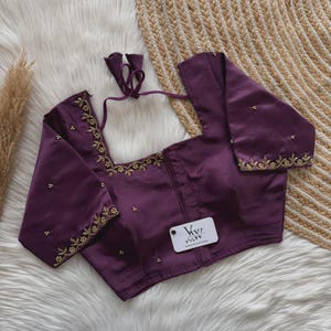 Pode incluir: Uma blusa roxa escura com decote quadrado e mangas curtas. A peça é adornada com bordados florais dourados ao longo do decote e das bordas das mangas, acentuados por pequenas contas espalhadas. Uma etiqueta branca com o texto VVI está anexada.