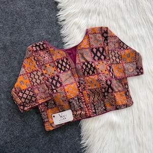 May include: A short-sleeved blouse featuring a patchwork design with various geometric and floral patterns in shades of orange, maroon, black, and grey. The garment has a V-neckline and a white rectangular tag with the text VVI printed in black.