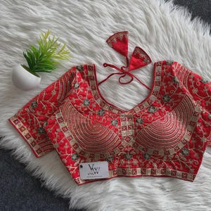 May include: A bright red blouse featuring intricate gold embroidery in floral and geometric patterns. The garment has a scoop neckline, short sleeves, and a back tie closure with two triangular tassels. A white tag with the text VVI is attached.