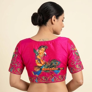 May include: A vibrant fuchsia short-sleeved blouse featuring a detailed back illustration of a figure holding a peacock feather and a flute. The garment includes intricate floral and paisley embroidery along the sleeves and bottom hemline.