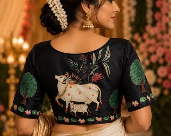 May include: A black short-sleeved blouse featuring a hand-painted design of a white cow and calf, surrounded by green trees and floral motifs. The garment has a round neckline and is paired with a cream-coloured fabric draped at the waist.