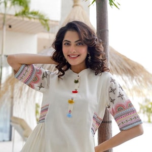 May include: White dress with colorful embroidered sleeves and accents. The dress features a mandarin collar and decorative tassels. The sleeves have a detailed pattern in pink, blue, and yellow. The dress is a light, flowing style.