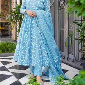 May include: A light blue dress and trousers set with a white geometric pattern. The dress has long sleeves and a V-neck. A matching scarf is draped over the shoulder. The outfit is paired with gold-colored flats. The outfit is made of a lightweight fabric.