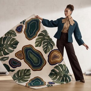 Può includere: Un tappeto rettangolare con sfondo crema presenta grandi foglie verdi di monstera e forme astratte nei toni del blu, marrone e verde. Il tappeto viene mostrato, mettendo in risalto il suo design e la sua consistenza.