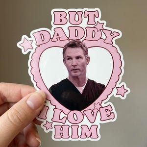 May include: A die-cut sticker featuring a pink heart-shaped frame with scalloped edges and small stars. Inside the heart is a black and white portrait of a man in a dark shirt. The text BUT DADDY I LOVE HIM is printed in pink block letters.