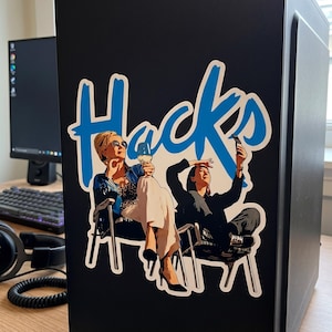 May include: A black computer tower featuring a large sticker with the blue text "Hacks" above an illustration of two people sitting in chairs. One person wears sunglasses and holds a glass, while the other holds a phone up to take a photo.