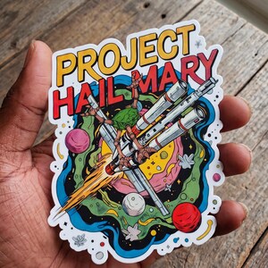May include: A die-cut sticker featuring the text PROJECT HAIL MARY in yellow and red letters. The illustration depicts a multi-limbed green creature piloting a silver spacecraft through a colorful space scene with planets, stars, and swirling blue and green cosmic clouds.