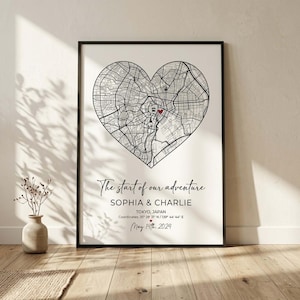 Puede incluir: Un estampado enmarcado con un mapa de Tokio en forma de corazón. El mapa en blanco y negro tiene un marcador rojo. El texto dice "The start of our adventure" con nombres y una fecha. El marco es negro.