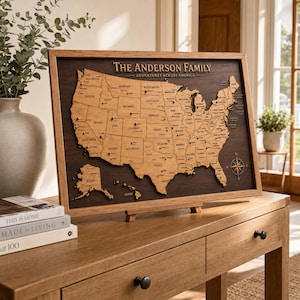 May include: A framed wooden map of the United States featuring the text The Anderson Family Adventures Across America. The map includes laser-cut state outlines, labeled cities, and small metal pins placed across the surface, set against a dark wood background.