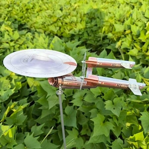 Könnte beinhalten: Ein silbernes und kupferfarbenes Modell eines Raumschiffs mit einer kreisförmigen Scheibe und zwei zylindrischen Abschnitten mit der Aufschrift "NCC-17". Das Modell ist auf einer dünnen Stange vor grünem Laubwerk montiert.
