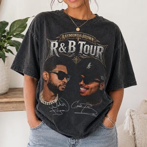 May include: A charcoal gray short-sleeve t-shirt featuring a graphic print that reads Raymond & Brown R&B Tour. The design includes portraits of two individuals wearing sunglasses, along with printed signatures below each face on the front.