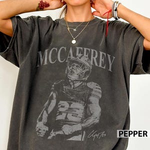 May include: A charcoal grey short-sleeve t-shirt featuring a distressed graphic of a football player in a helmet and jersey with the number 23. The text MCCAFFREY is printed in a large arched font above the player, with a signature below.