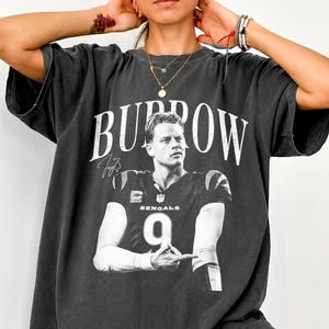 May include: A charcoal grey short-sleeve t-shirt featuring a large black and white graphic of a football player wearing a jersey with the number 9. The word BURROW is printed in large white capital letters above the player, with a signature below.