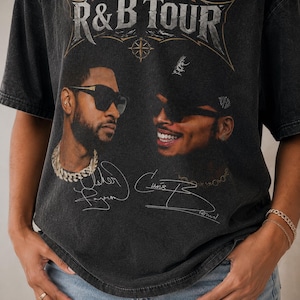 May include: A charcoal gray short-sleeve t-shirt featuring a graphic print with the text R&B TOUR at the top. The design includes portraits of two men wearing sunglasses, accompanied by printed signatures below each face on the front of the shirt.