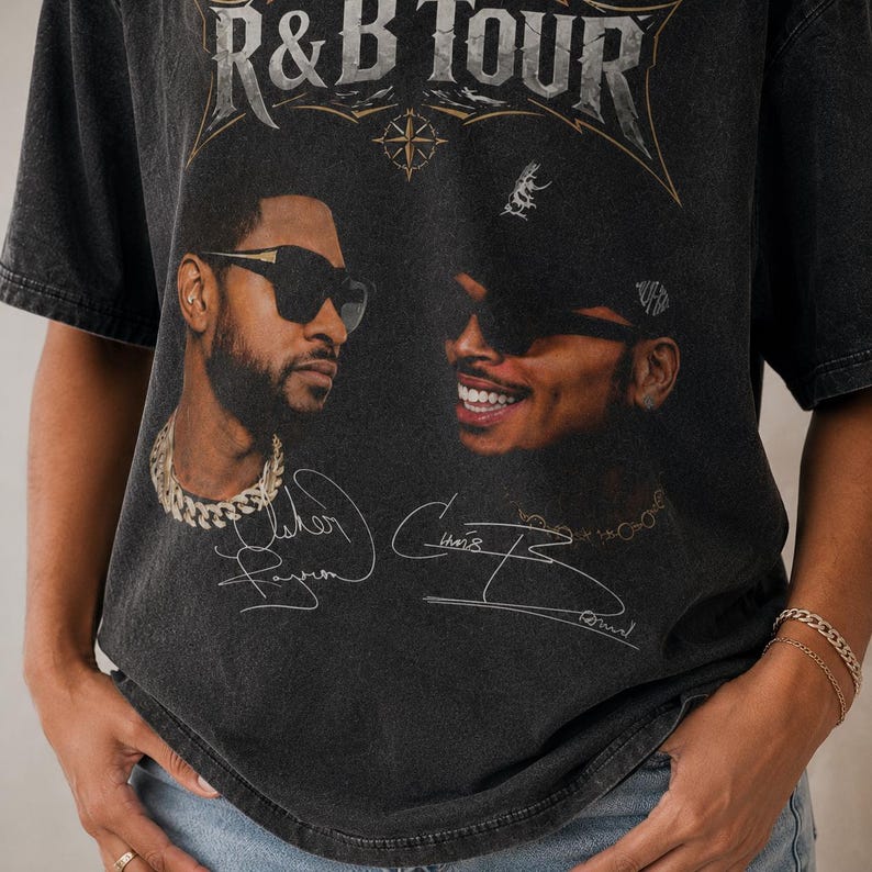 May include: A charcoal gray short-sleeve t-shirt featuring a graphic print with the text R&B TOUR at the top. The design includes portraits of two men wearing sunglasses, accompanied by printed signatures below each face on the front of the shirt.