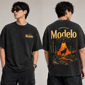 Op de afbeelding: Zwart T-shirt met korte mouwen met het woord "Modelo" in geel op de voorkant. De achterkant toont een woestijnscène met een stier, cactussen en een UFO, met de woorden "CERVEZA Modelo".