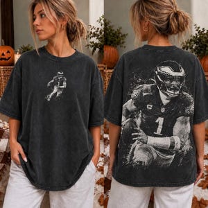 Può includere: Una t-shirt oversize grigio antracite con una piccola grafica di un giocatore di football sul petto a sinistra e una grande grafica in bianco e nero dall'aspetto vissuto di un giocatore con il numero 1 sul retro del capo.