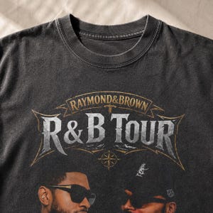 May include: A charcoal gray crew neck t-shirt featuring a graphic print with the text RAYMOND & BROWN R&B TOUR in a weathered, metallic-style font. The design includes a compass rose and two individuals wearing sunglasses at the bottom.