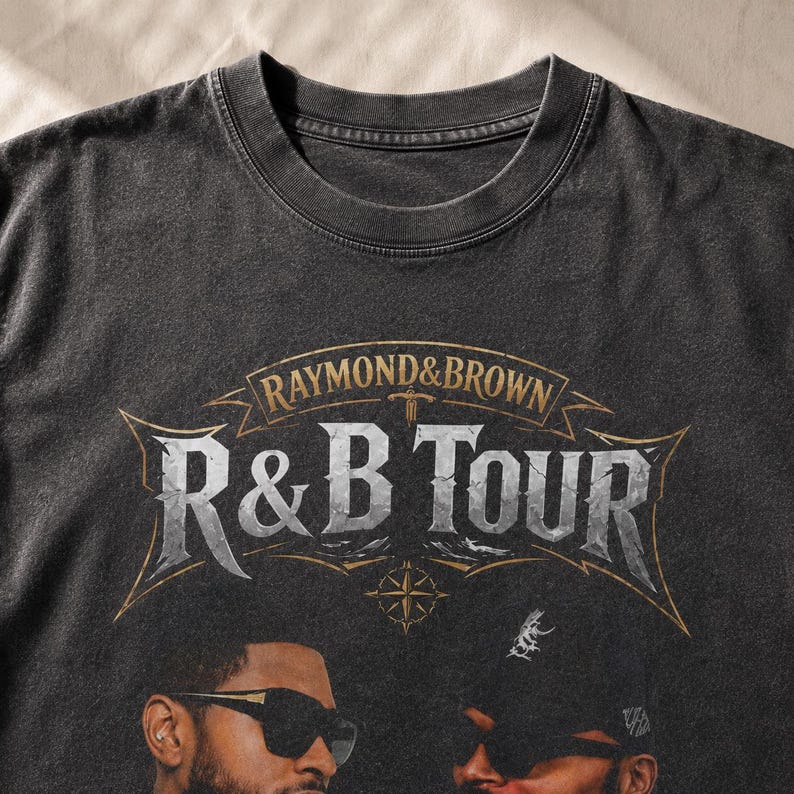 May include: A charcoal gray crew neck t-shirt featuring a graphic print with the text RAYMOND & BROWN R&B TOUR in a weathered, metallic-style font. The design includes a compass rose and two individuals wearing sunglasses at the bottom.