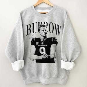 May include: A heather grey crewneck sweatshirt featuring a black and white graphic of a football player in a jersey with the number 9 and the word BENGALS. The name BURROW is printed in large black letters above the player, with a signature below.