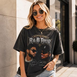 May include: A charcoal grey short-sleeve t-shirt featuring a graphic print with the text "RAYMOND & BROWN R&B TOUR". The design includes portraits of two individuals wearing sunglasses, along with printed signatures below each portrait.