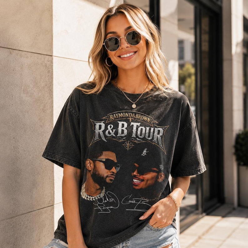 May include: A charcoal grey short-sleeve t-shirt featuring a graphic print with the text "RAYMOND & BROWN R&B TOUR". The design includes portraits of two individuals wearing sunglasses, along with printed signatures below each portrait.