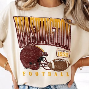 May include: A cream-coloured t-shirt featuring a distressed graphic with the word WASHINGTON in large yellow and maroon letters, a maroon football helmet, a brown football, and a rectangular badge with the text EST. 1932 above the word FOOTBALL.