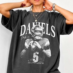 May include: A charcoal gray short-sleeve t-shirt featuring a white graphic print of a football player wearing a helmet and holding a football. The text DANIELS is printed in large white letters above the player, with the number 5 below.