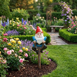 May include: A garden gnome figurine featuring a red pointed hat, a long white beard, and a blue shirt, mounted on a brown wooden stake. The gnome is positioned upright in a garden bed filled with pink, yellow, and purple flowers.