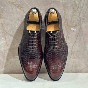 May include: A pair of dark brown oxford shoes featuring a textured snakeskin pattern, a pointed toe, and a five-eyelet lace-up closure. Each shoe contains a light-colored wooden shoe tree insert to maintain the shape of the footwear.