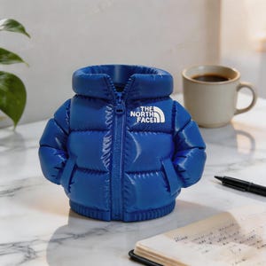 May include: A bright blue miniature puffer jacket-shaped desk tidy. The jacket has a zipper and the white "THE NORTH FACE" logo. The desk accessory is on a marble surface, with a coffee mug and pen in the background.