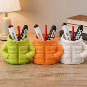 Puede incluir: Tres portalápices con forma de chaquetas acolchadas en verde lima, naranja y blanco. Cada chaqueta contiene bolígrafos y rotuladores con detalles en rojo y negro. Los portalápices están sobre una superficie de madera.