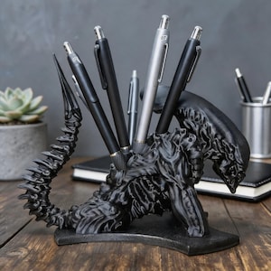 May include: A black pen holder in the shape of a creature, holding several pens. The pen holder is made of a dark material and has a detailed, textured design. The pens are black and silver, and are of various styles.