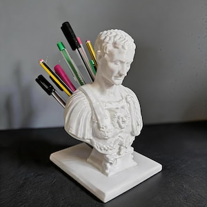 May include: A white ceramic bust of a Roman emperor, used as a pen holder. The bust is on a square base and holds several pens of various colors. The pens are black, green, pink, and yellow. The bust is a decorative desk accessory.