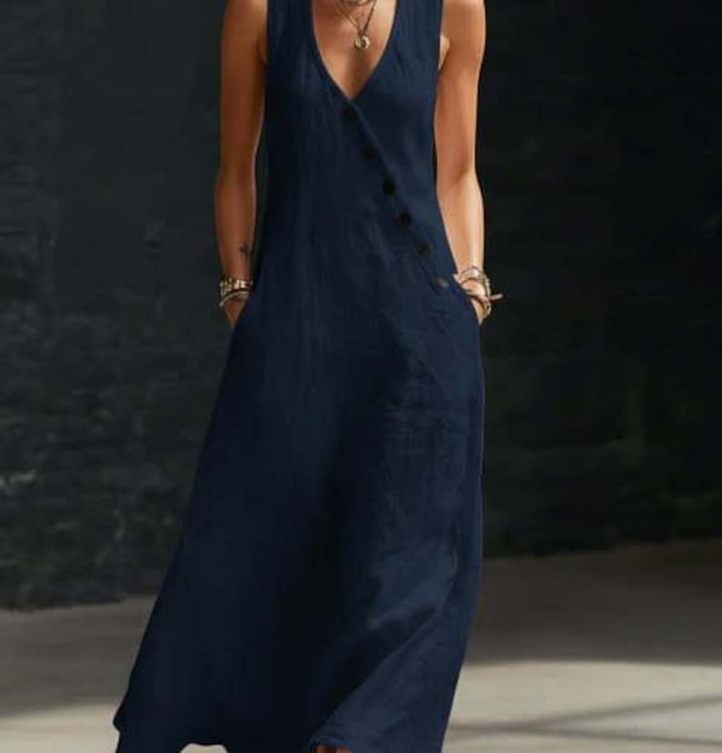 May include: A sleeveless, navy blue jumpsuit with a V-neckline and a button closure. The jumpsuit features two pockets and a flowing, wide-leg design. The fabric appears to be a lightweight linen or cotton blend.