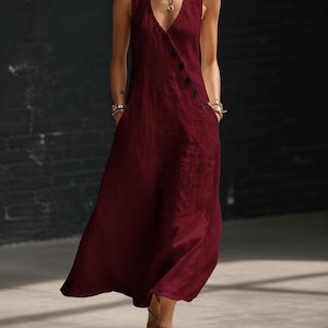 May include: A sleeveless, burgundy-colored dress with a V-neckline and a button closure. The dress has pockets and a flowing, floor-length design. The image also shows the text "AveloraStyle" in the upper left corner.