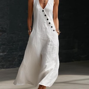 May include: A sleeveless white jumpsuit with a V-neckline and wide legs. The jumpsuit features a row of black buttons down the front and pockets. The fabric appears to be a lightweight linen or cotton blend, suitable for warm weather.