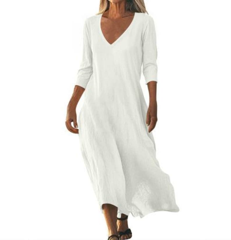 May include: A flowing, off-white dress with a V-neck and three-quarter sleeves. The dress has a relaxed fit and falls to the mid-calf. The fabric appears to be lightweight and textured, suitable for warm weather. The model is wearing black sandals.