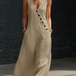 May include: A sleeveless, beige jumpsuit with a V-neckline and a row of black buttons down the side. The jumpsuit has pockets and a wide-leg design, made of a lightweight fabric.