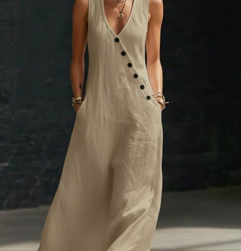 May include: A sleeveless, beige jumpsuit with a V-neckline and a row of black buttons down the side. The jumpsuit has pockets and a wide-leg design, made of a lightweight fabric.