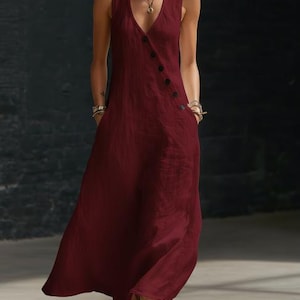May include: A sleeveless, burgundy maxi dress with a V-neckline and a button-up closure. The dress features two pockets and a flowing, floor-length skirt. The fabric appears to be a lightweight linen or cotton blend, suitable for warm weather.